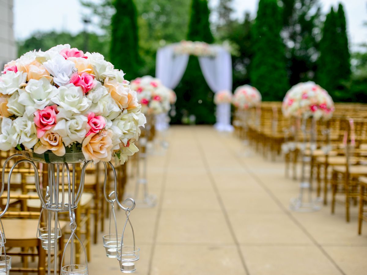 An outdoor wedding setup with rows of chairs, floral arrangements featuring white and pink flowers, and an archway decorated with white fabric and flowers.