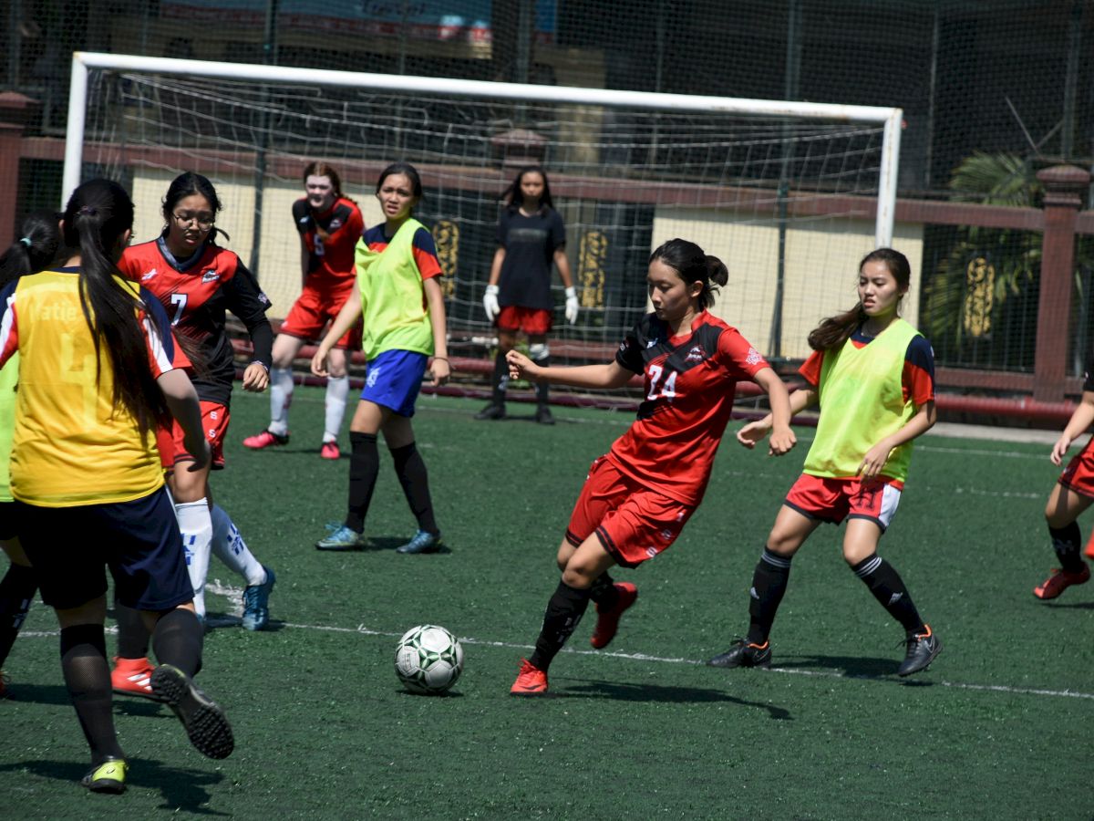 A group of female soccer players in red and black uniforms compete for the ball on a field, with a goal and net in the background.