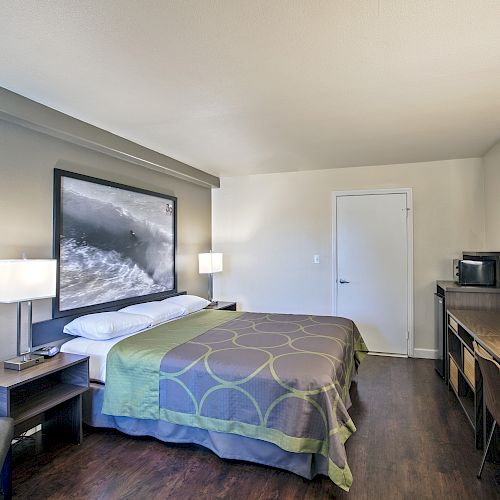 A clean, modern hotel room with a large bed, two bedside tables with lamps, a desk area with a chair, a TV, and a microwave on top of the desk.