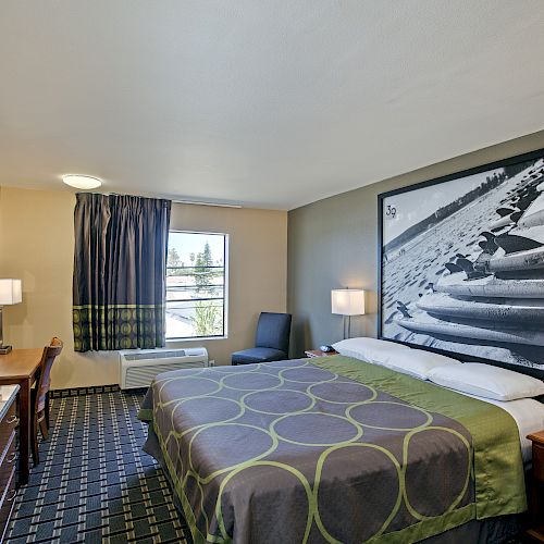 The image shows a hotel room with a king-sized bed, two bedside tables with lamps, a desk with a chair, a TV, and a window with curtains.