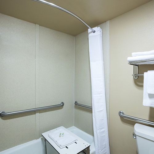 A bathroom with a shower, white curtain, grab bars, a folding seat, wall shelves with towels, and a toilet with a towel rack above it.