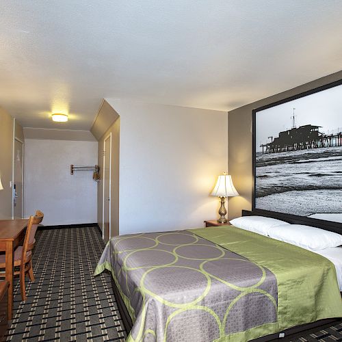 A hotel room with a bed, two lamps, a desk with a chair, a TV, and a large black-and-white picture above the bed. The floor has a patterned carpet.