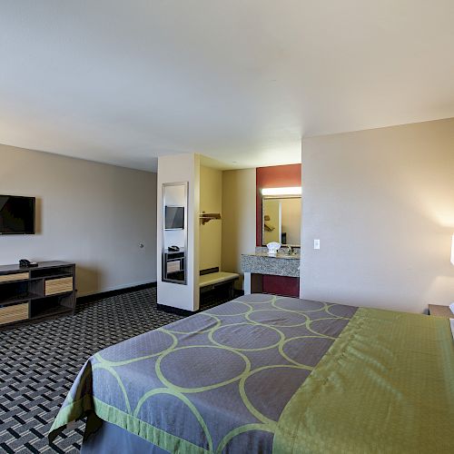 The image shows a hotel room with a bed, desk, TV, lamp, and a vanity area with a sink and mirror. The bedding is green with circular patterns.