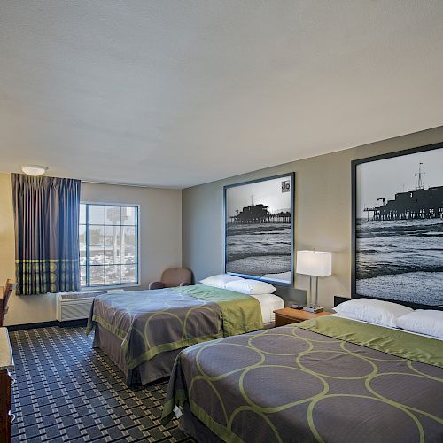 A hotel room with two double beds, bedside lamps, wall art of piers, a desk, TV, and a window with curtains letting in natural light.