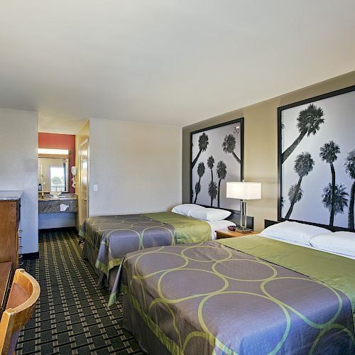 The image shows a hotel room with two double beds, a flat-screen TV, and artwork featuring palm trees on the walls.