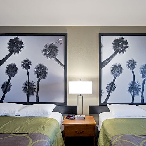 The image shows two beds with green covers, separated by a nightstand with a lamp, and two large black-and-white palm tree pictures behind the beds.