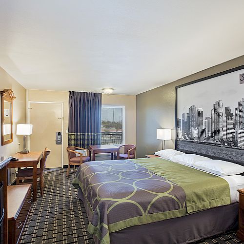 This image shows a tidy hotel room with a bed, two lamps, a TV, a desk with a chair, a small table with two chairs, and wall art of a cityscape.