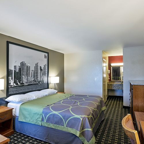 A hotel room with a bed, a framed cityscape, nightstands, a TV, a desk, a chair, and an open vanity area leading to the bathroom at the side.