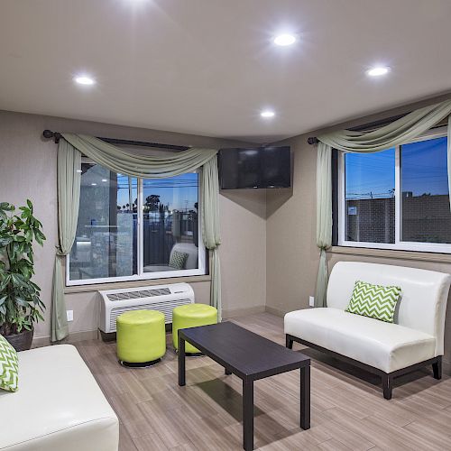 A modern lounge with white sofas, green pillows, a dark table, green ottomans, large windows with drapes, a wall-mounted TV, and potted plant.