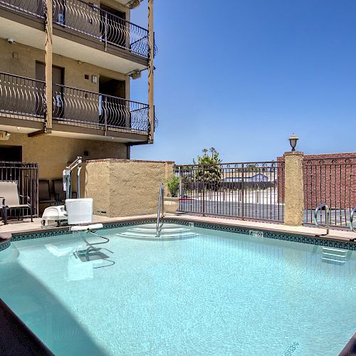 An outdoor swimming pool with a lift for disabled access, surrounded by chairs and a building with balconies, under a clear blue sky.