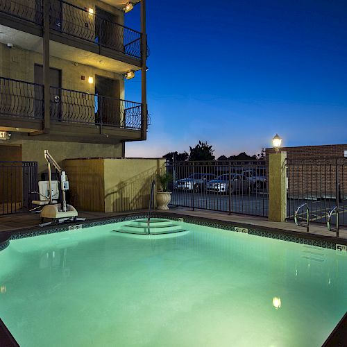 A lit pool area at night with an adjacent building, parking area, and seating. The scene has a serene ambiance with subtle lighting.