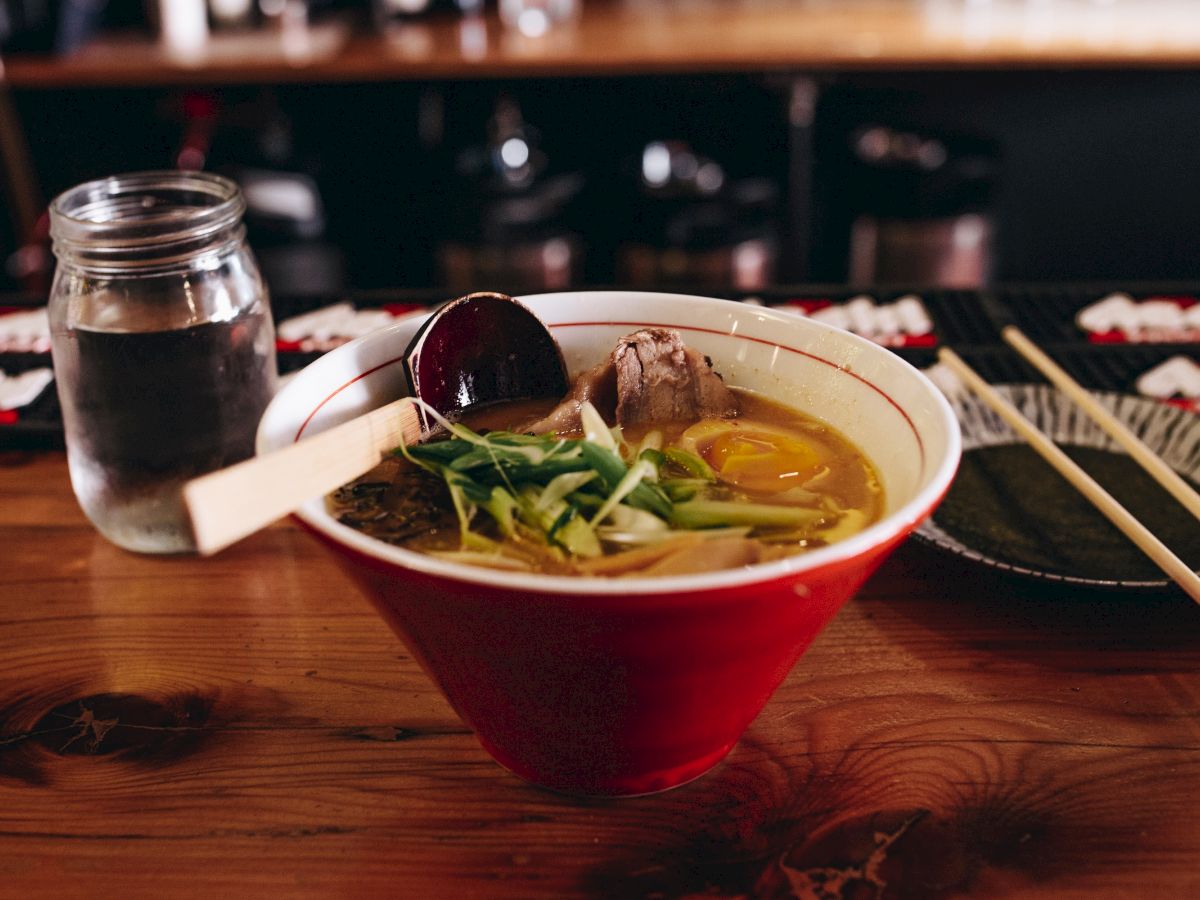 This image features a bowl of ramen with a jar of water, chopsticks, and a spoon on a wooden table in a restaurant setting.