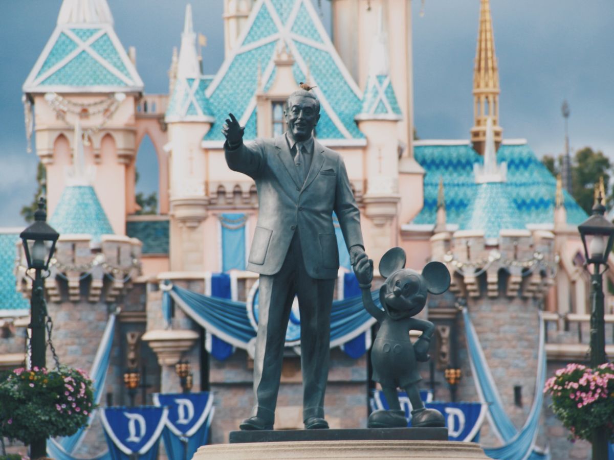 The image shows a statue of a man and a mouse character in front of a castle with blue rooftops, likely in a themed amusement park.