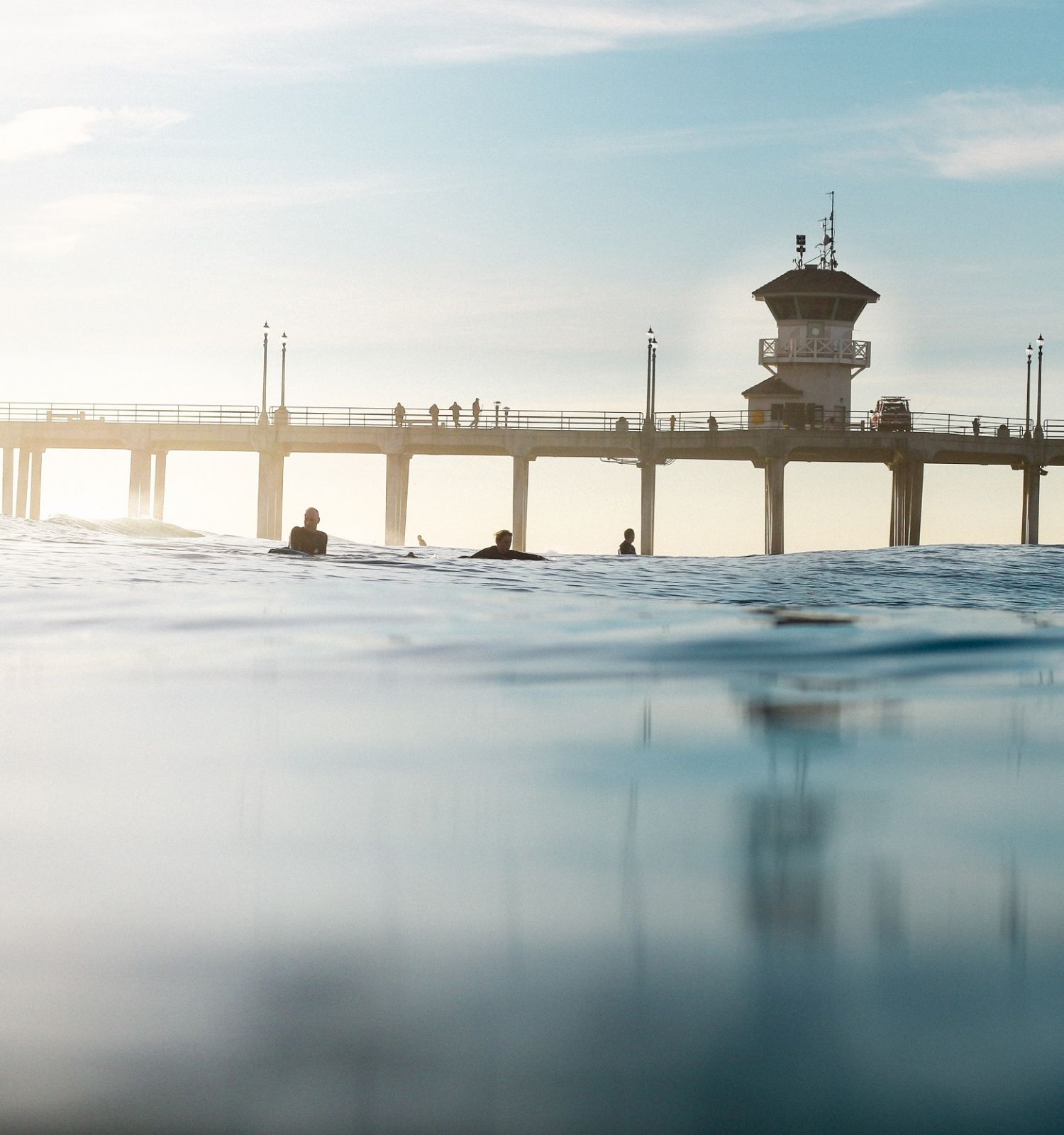 The image shows a calm ocean with a few people in the water and a long pier in the background under a clear sky.