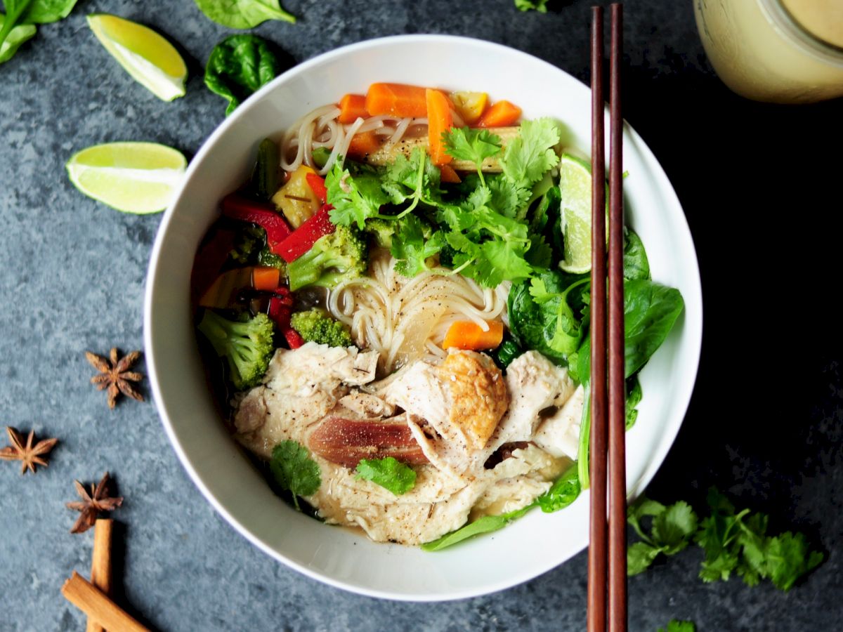 The image shows a bowl of noodle soup with vegetables, herbs, and possibly chicken, accompanied by lime wedges and spices on a dark surface.