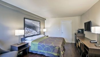 A tidy hotel room with a queen-sized bed, modern decor, a flat-screen TV, a desk, lamps, and a chair, all against neutral-colored walls.