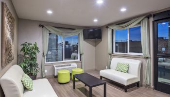 A modern living room with white sofas, green pillows, a black coffee table, green stools, wall art, plants, and a wall-mounted TV, is shown.