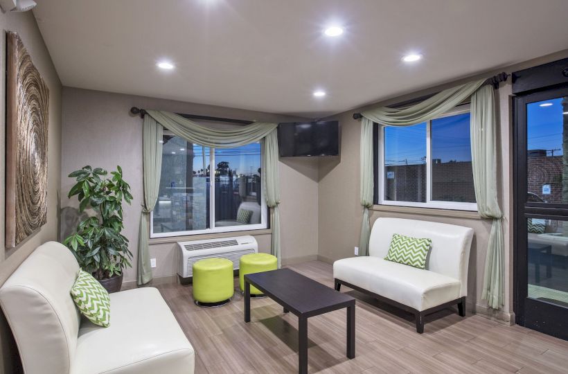 A modern living room with white sofas, green pillows, a black coffee table, green stools, wall art, plants, and a wall-mounted TV, is shown.