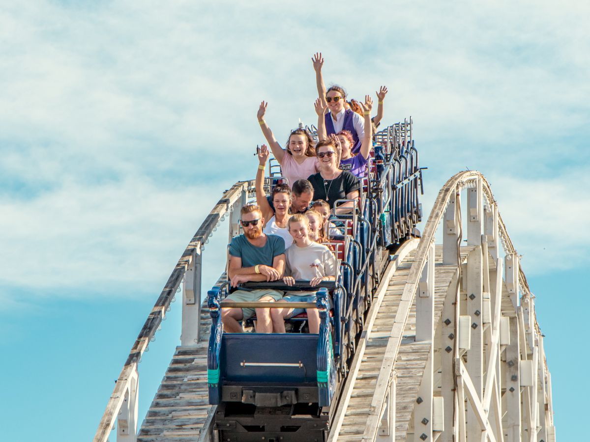 A group of people on a roller coaster, raising their hands in excitement as they descend a steep drop under a bright blue sky.