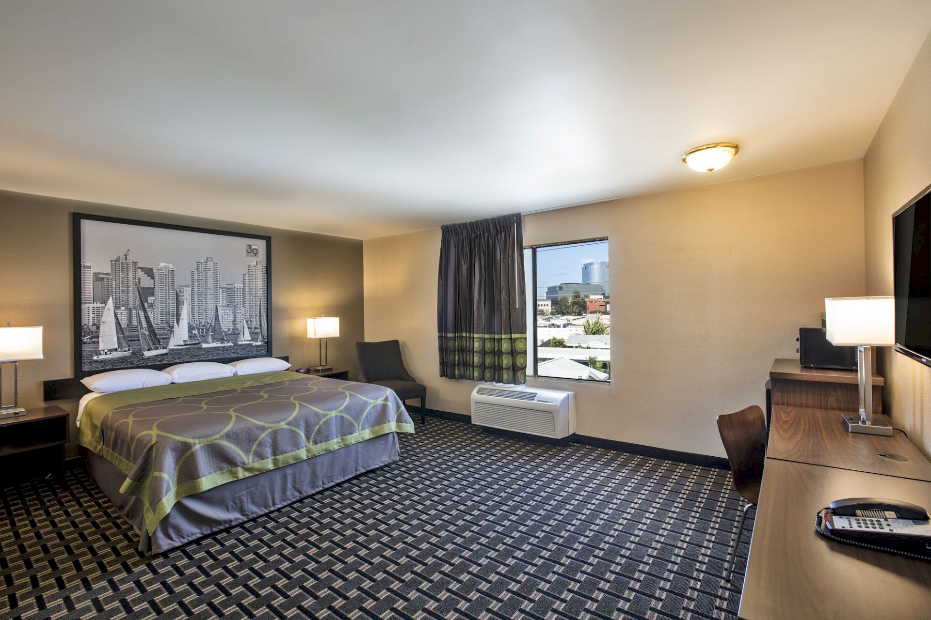 A hotel room with a king-size bed, artwork above the bed, two nightstands with lamps, a window, an air conditioner, a desk, and a chair.