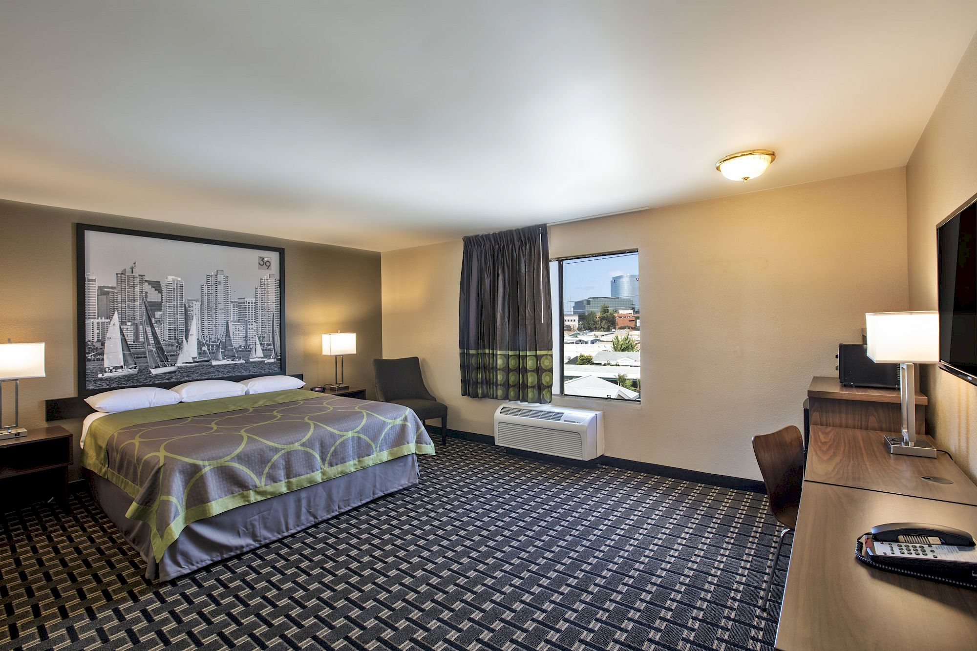 A hotel room with a king-size bed, artwork above the bed, two nightstands with lamps, a window, an air conditioner, a desk, and a chair.