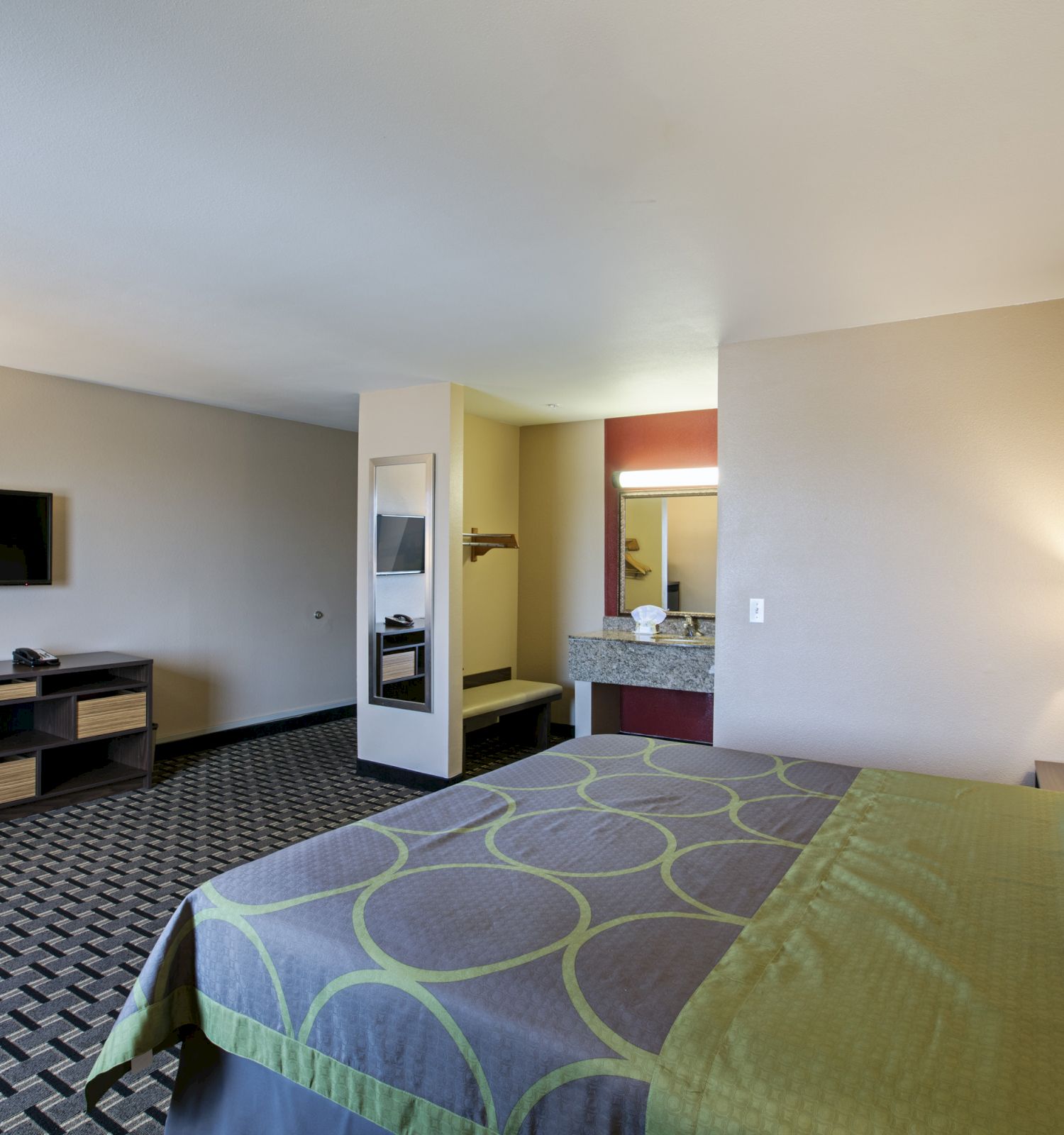 The image shows a hotel room with a bed, TV, desk, chair, and a sink area in the background. The decor is modern and simple.