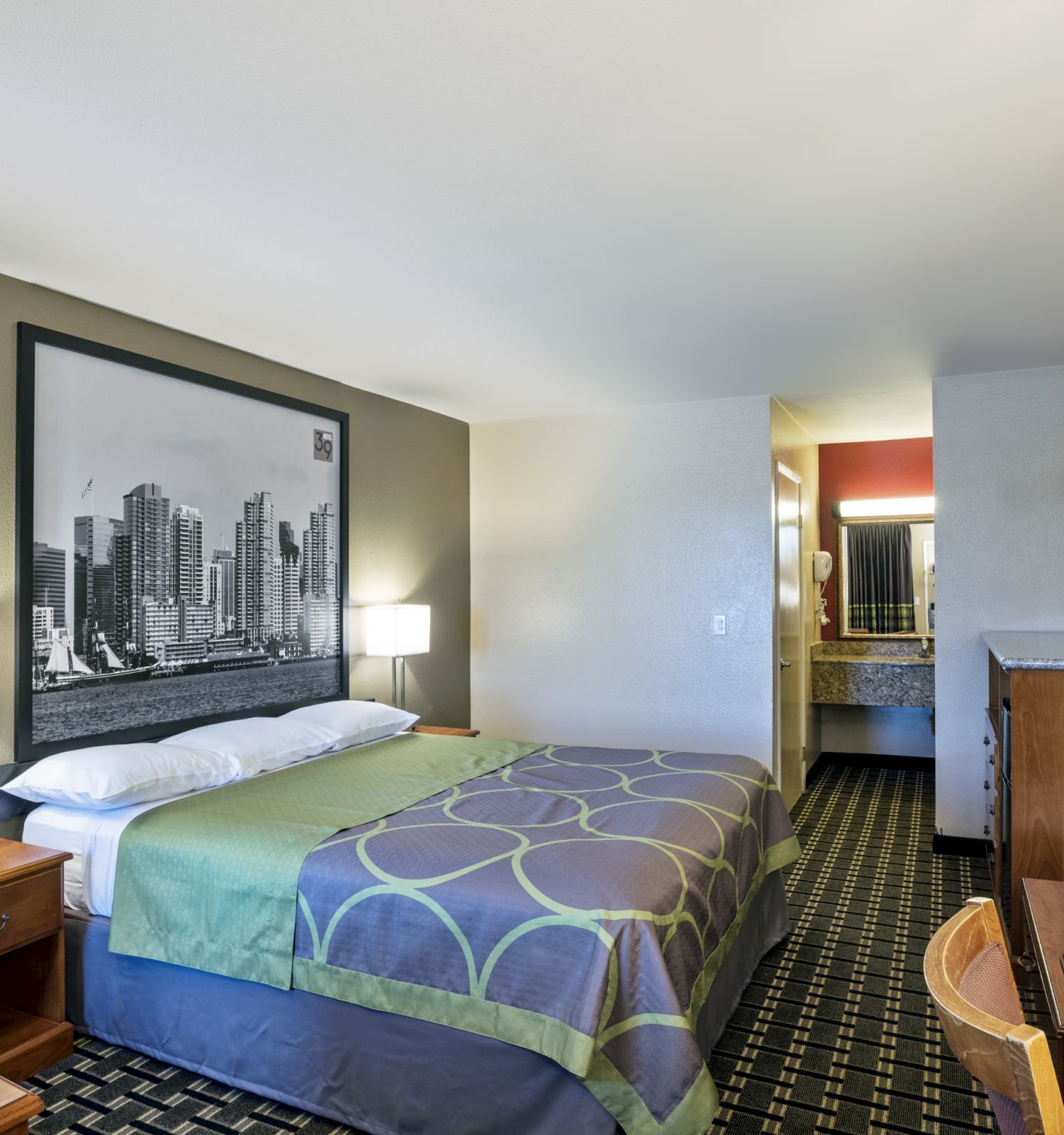 The image shows a hotel room with a double bed, lamps on nightstands, a cityscape picture, a TV, wooden furniture, and a sink area in the background.